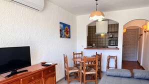 A compact living space with a TV, wooden dining table, chairs, a sofa, a kitchen area with a microwave, and a door with a patterned design. - 0167-POBLAT TIPIC Studio near the beach (Empuriabrava)
