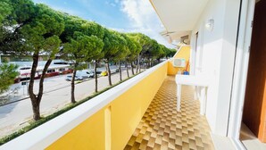 A balcony with a yellow wall, a patterned floor, and a view of a street with trees and parked cars. - Cozy apartment with large balcony (Bibione)