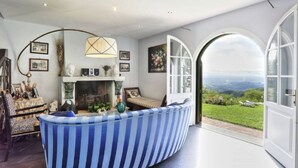 A living room with a fireplace, a striped sofa, and a view of the countryside through large windows. - Elegant Villa near Lucca with big and panoramic garden and private pool (Capannori)