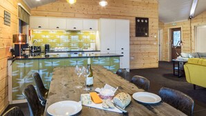 Un interior de madera con una mesa de comedor preparada para dos comensales, una zona de cocina y un sofá amarillo.