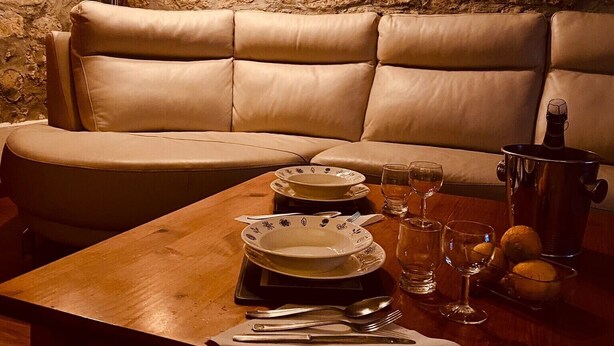 Un coin repas accueillant avec un mur en pierre, un banc rembourré, une table en bois dressée pour deux personnes et une bouteille de vin.
