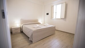 A bedroom with a bed, bedside tables, a window, and a wall-mounted light switch. - Villa Barletta apartment in villa ground floor n.2 (Marina di Acate)