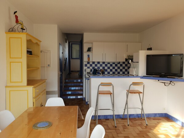 Cocina compacta con salpicadero a cuadros, un televisor montado sobre la encimera y comedor con mesa de madera y sillas blancas.