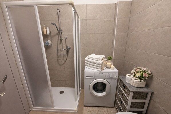 A bathroom with a walk-in shower, a washing machine, and a small shelf with toiletries. - Casa Gina Giardino Holideal Apartment (Tremosine)