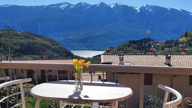 A balcony with a table and chairs, a vase with yellow flowers, and a view of mountains and a lake.