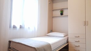 A single bed with a mattress and pillows, a small shelf with a potted plant, and a wardrobe with multiple drawers.