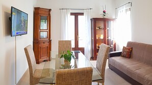 A living room with a glass-top dining table, wicker chairs, a beige sofa, and a television mounted on the wall.