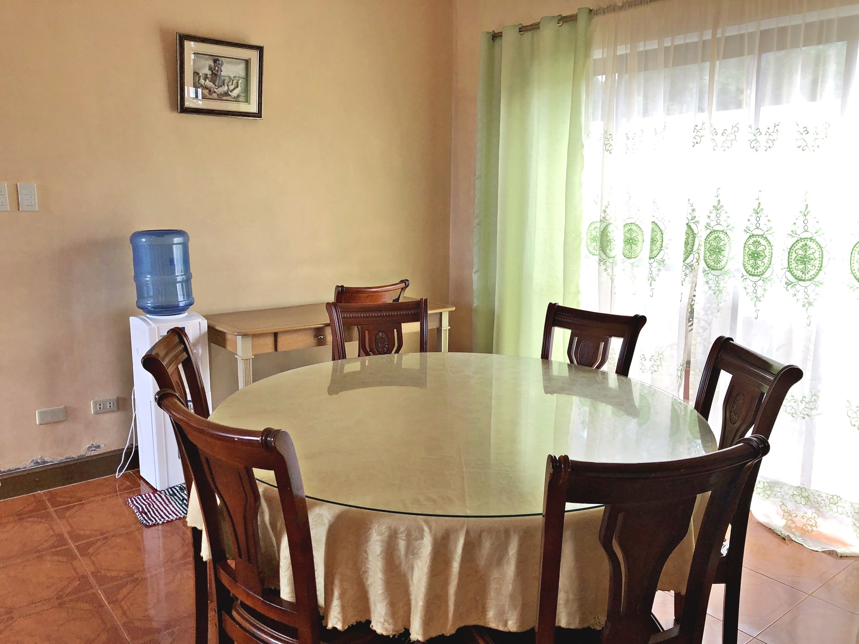 A round glass table with a beige tablecloth, surrounded by wooden chairs. A water dispenser is on the left, and a picture hangs on the wall.