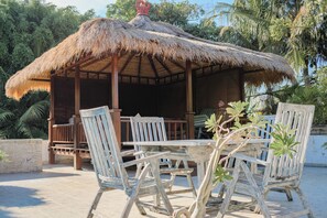 A wooden gazebo with a thatched roof, surrounded by white chairs and a table, set against a backdrop of lush greenery. - Fabulous villa right on the beach with large pool, tropical garden & staff (Dencarik/Lovina)