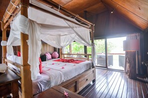 A wooden four-poster bed with white bedding and red accents, a wooden bench, and a view of greenery outside. - Fabulous villa right on the beach with large pool, tropical garden & staff (Dencarik/Lovina)
