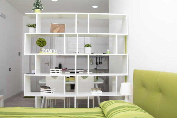 A modern living room with a white shelving unit, a green couch, and a kitchen area in the background.