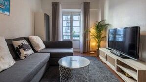 Una sala de estar moderna con un sofá gris, una mesa de centro redonda, un televisor sobre un mueble de madera y un ventanal con cortinas.