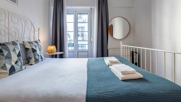 Un dormitorio ordenado con cama, almohadas, una lámpara de noche y vistas al exterior por la ventana.