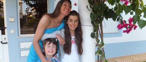 A family of three standing in front of a building with a blue exterior and a white column.