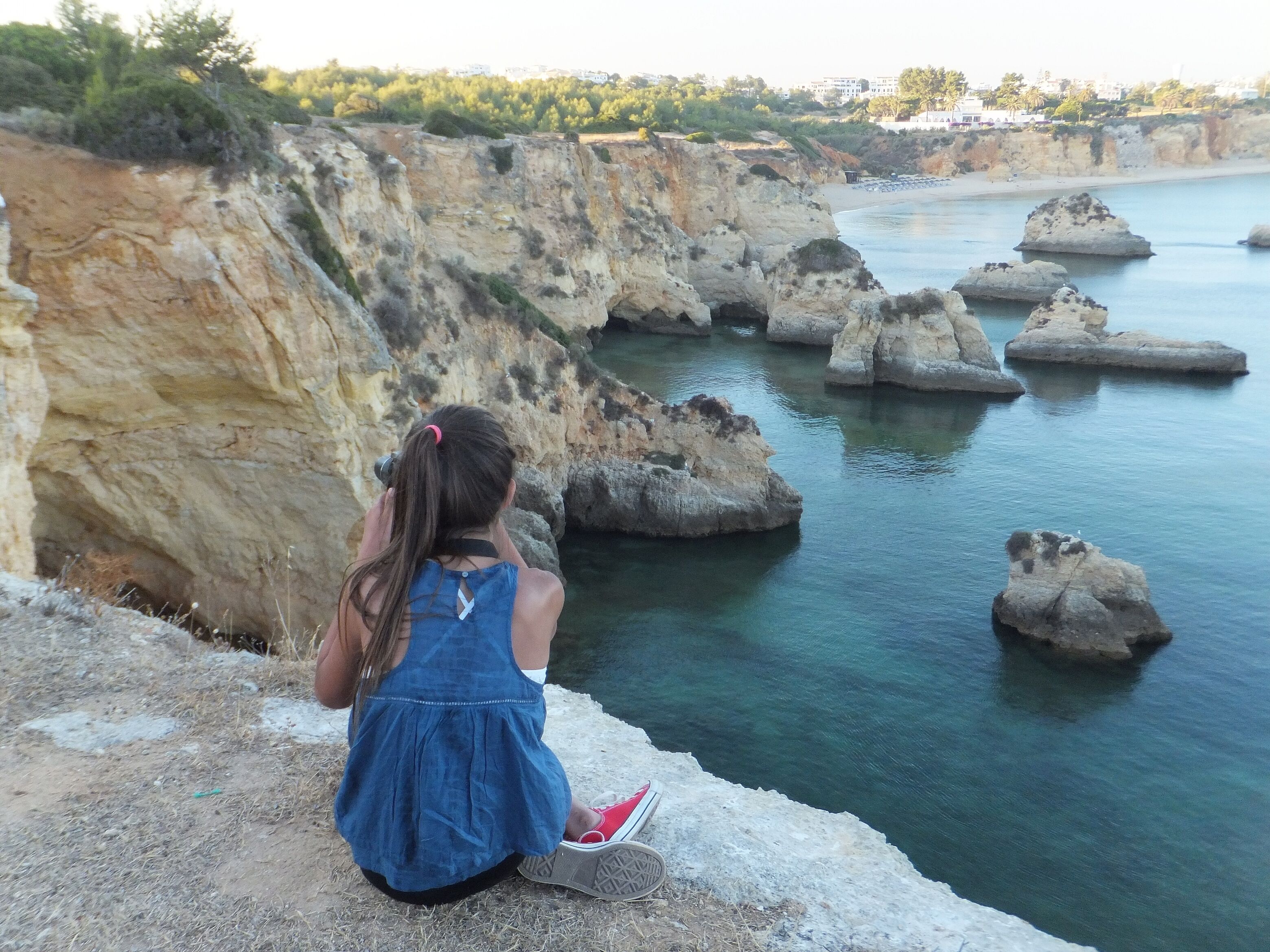 A person sitting on a cliff overlooking a coastal area with rocky formations and calm water.