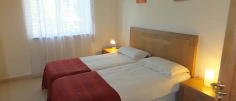Una cama doble con dos mantas rojas, un cabecero de madera, mesitas de noche con lámparas, una ventana con cortinas traslúcidas y dos obras de arte colgadas en la pared.