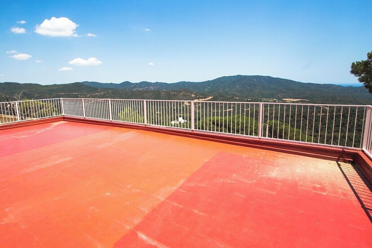 Une terrasse aux tuiles rouges, agrémentée d’une balustrade blanche, d’où l’on peut admirer des collines verdoyantes et un ciel dégagé.