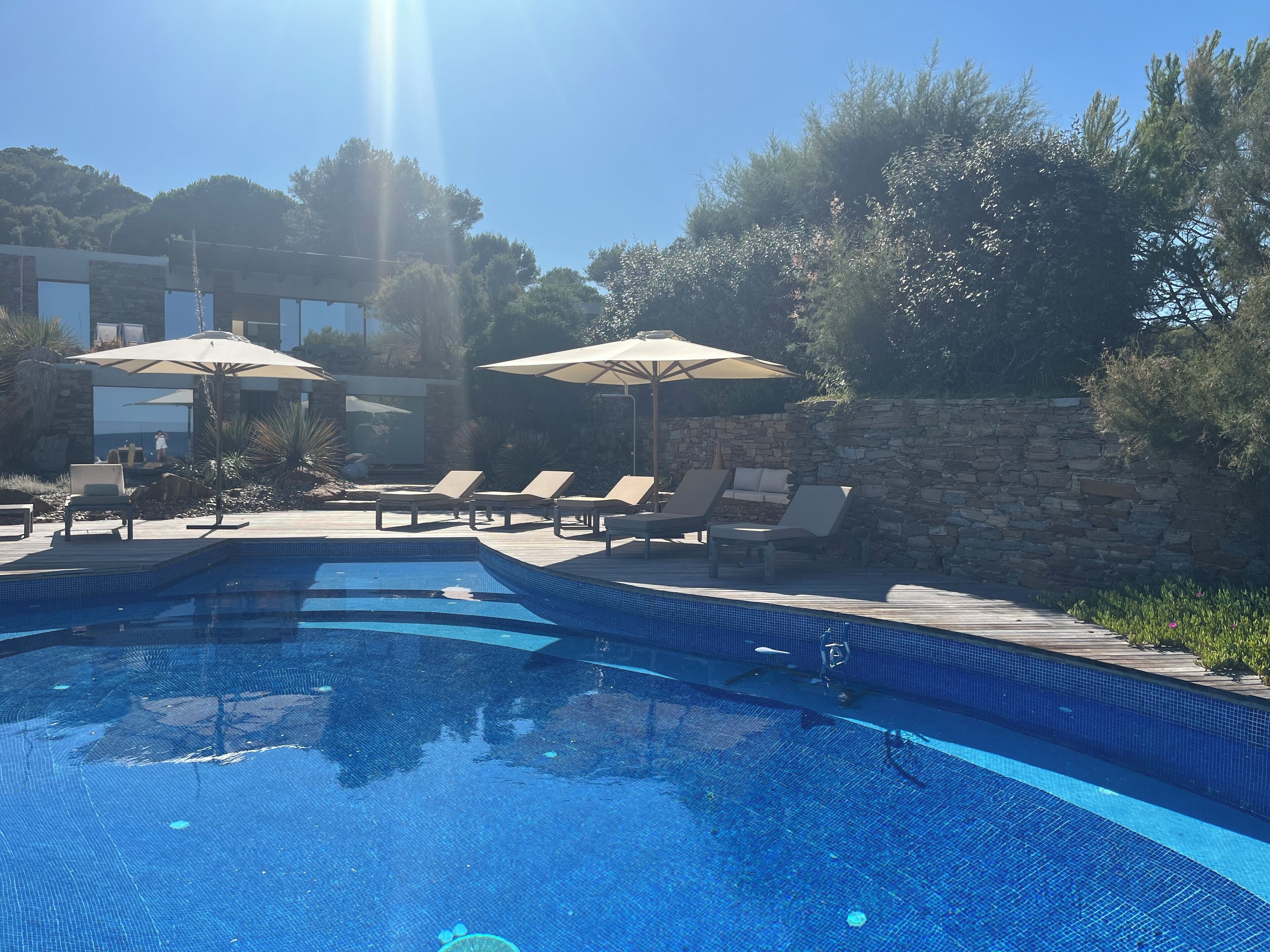 Ein Swimmingpool mit Liegestühlen und Sonnenschirmen, umgeben von Steinmauern und viel Grün.
