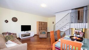 Una sala de estar con sofá, televisor, mueble de entretenimiento de madera, mesa de comedor con sillas y una escalera.