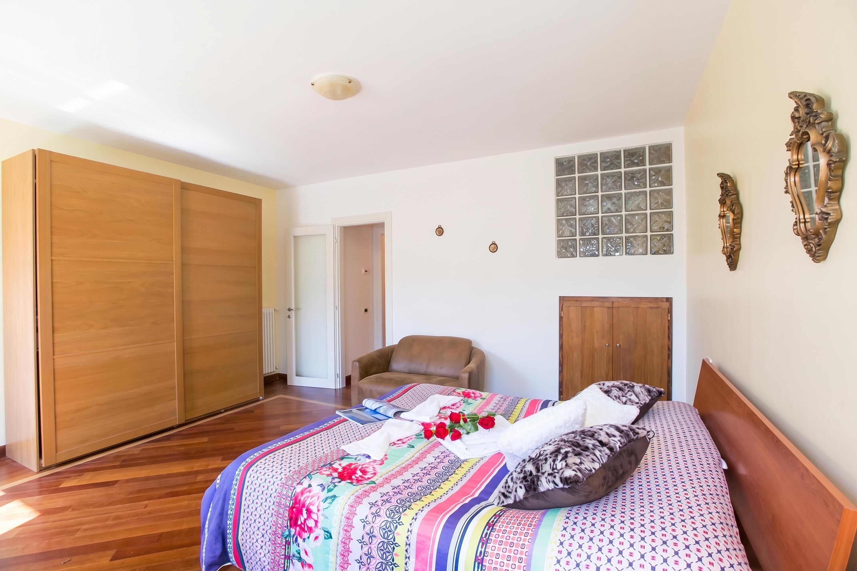 A bedroom with a large bed, a wooden wardrobe, a chair, and a window with decorative glass.