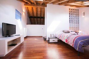 Une chambre moderne avec une télévision, un plancher en bois, un lit recouvert d’une courtepointe à motifs et une fenêtre avec des stores.
