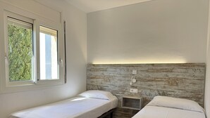 Una habitación con dos camas individuales, cabecera de madera, mesita de noche pequeña y ventana con vista a árboles.