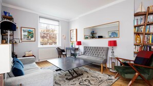 A living room with a grey sofa, a coffee table, a red armchair, a bookshelf, and a large window.