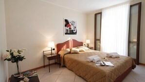A hotel room with a bed, two bedside tables with lamps, a nightstand with a vase of flowers, a large window with curtains, and a painting on the wall. - Residence Eden Magnolia 14, Argegno, Italy (Argegno)