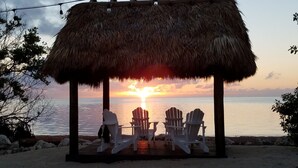 A thatched-roof structure with two white chairs and a table, overlooking a calm sea at sunset. - Key Largo Oceanfront Escape: Pools, Beach, Fishing Pier, No Steps ! 🏝️ (Key Largo)
