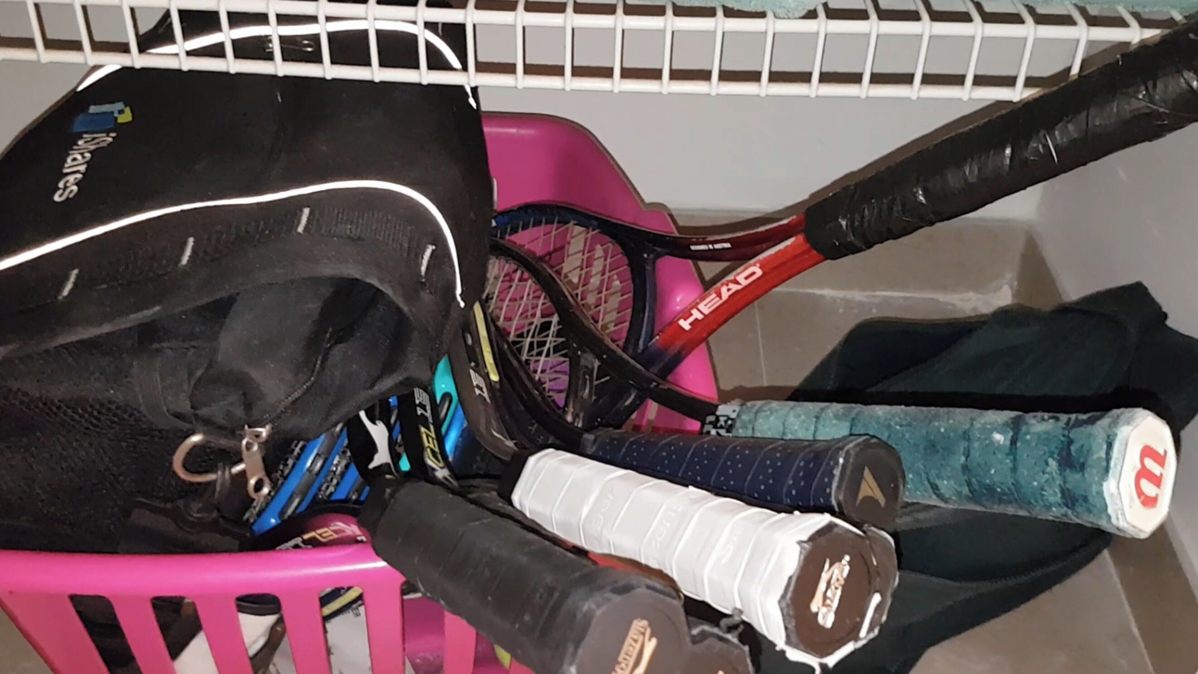 A pink basket containing a black bag, a red and black tennis racket, and a blue and white tennis racket.