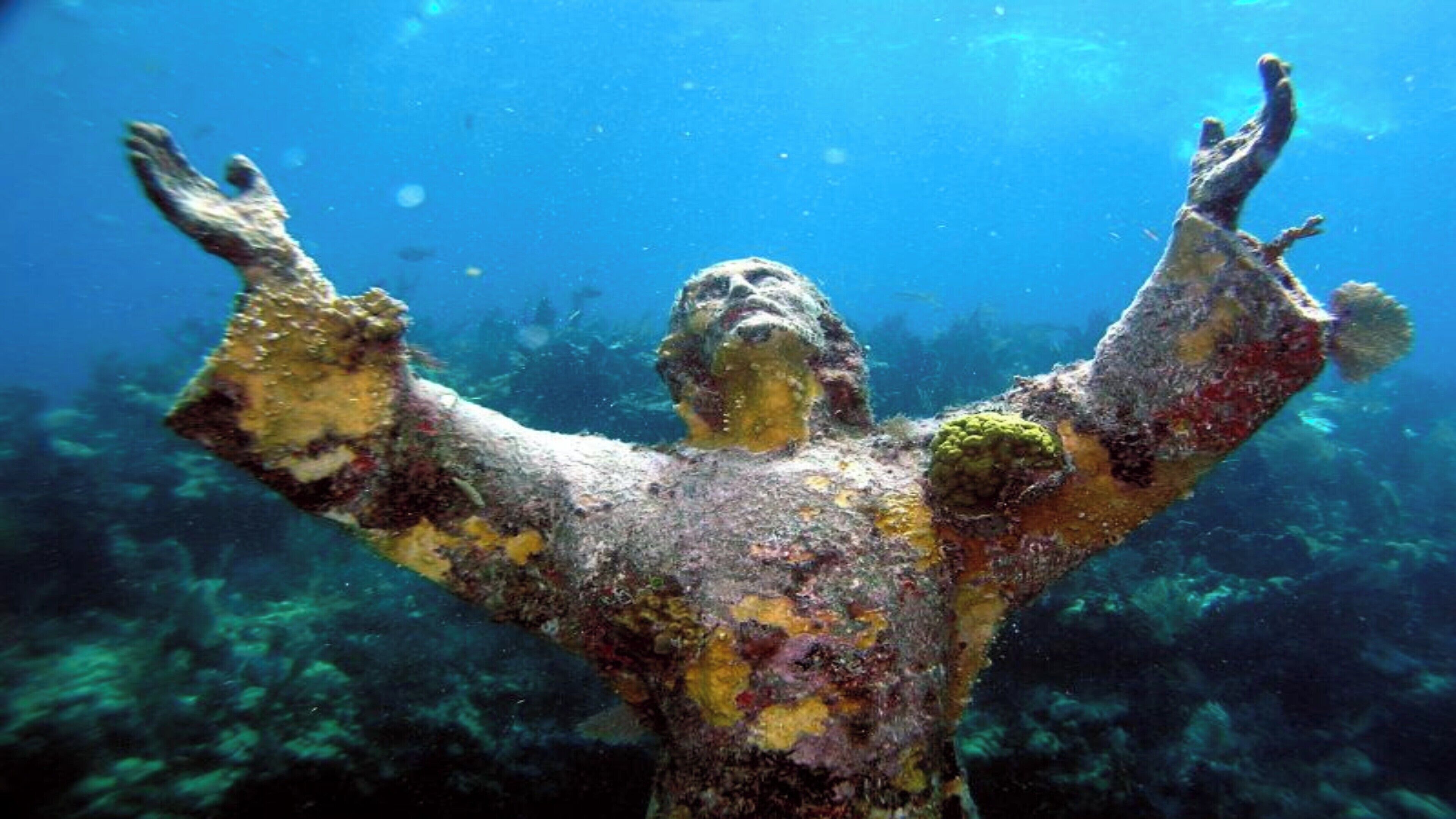 Una estàtua amb els braços estesos, parcialment coberta per creixements marins, sota l'aigua.