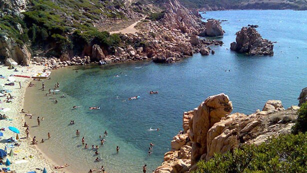 Ein Strand mit klarem Wasser, Felsen und badenden Personen.