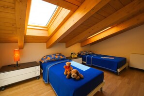 A room with a sloped ceiling, wooden beams, a skylight, two beds with blue bedding, a bedside table with a lamp, and a teddy bear on one bed. - Residence Eden Azalea 3, Argegno, Italy (Argegno)