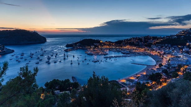 Una cittadina costiera al crepuscolo con un porto pieno di barche, un faro e edifici lungo la riva.
