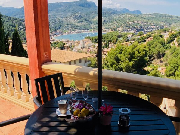 Un balcón con una mesa lista para dos personas, con vista a un paisaje pintoresco que incluye montañas y una masa de agua.