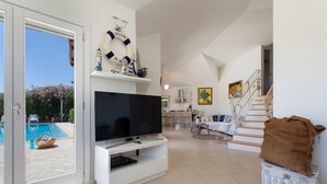 Een ruime woonkamer met een groot flatscreentelevisie, een rieten mand en een zwembad zichtbaar door glazen deuren.
