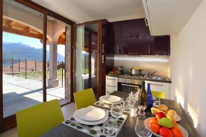 A dining area with a table set for two, a kitchen with modern appliances, and a view of mountains through a glass door. - Parco dei Cervi Terrazzo, Plesio, Italy (Plesio)