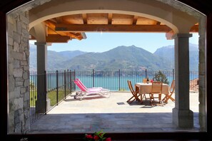 A balcony with a stone wall, a wooden pergola, a table with chairs, and a view of mountains and a lake.