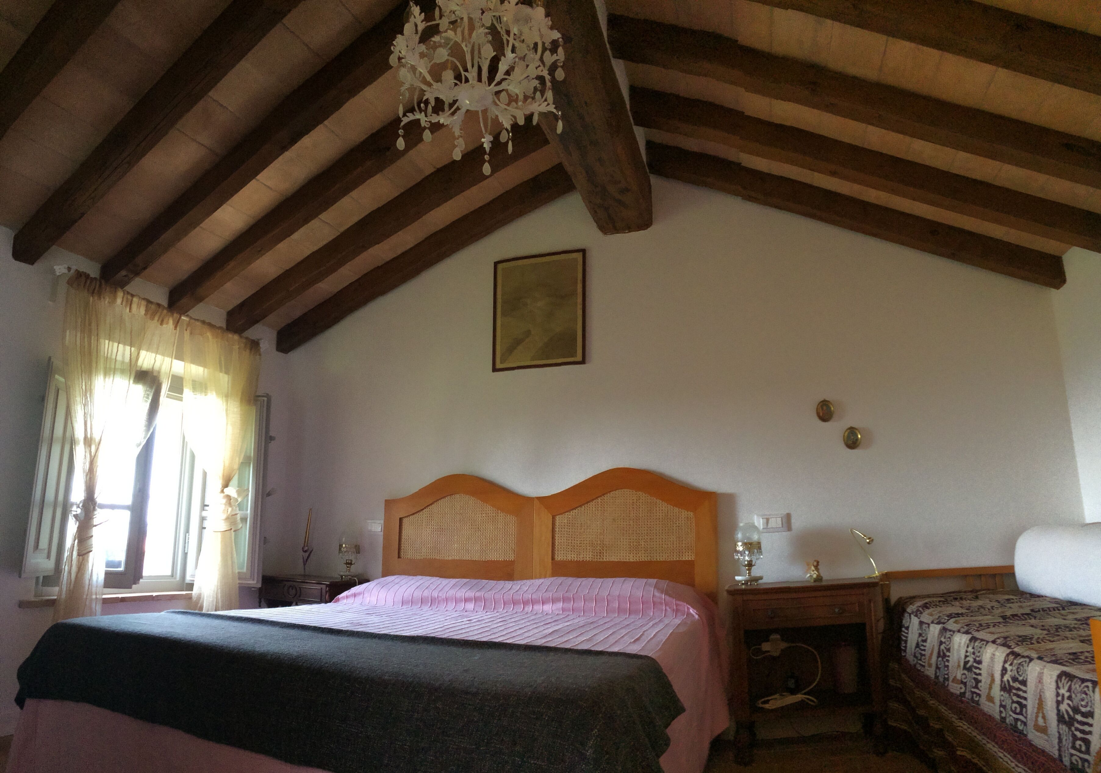 A bedroom with a wooden ceiling, a bed with a headboard, a bedside table, a lamp, and a window with curtains.