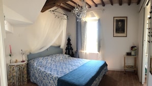Una camera da letto con un letto, un lampadario, una finestra con tende, un comodino con un vaso e un quadro appeso al muro.