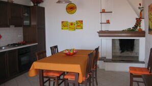 Un comedor con una mesa dispuesta para seis personas, rodeada de sillas de madera con cojines naranja. Se ve una chimenea al fondo, y hay tres cuadros en la pared sobre la chimenea.