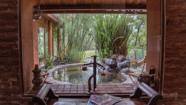 A rustic bathroom with a stone wall, a wooden bench, a small hot tub, and a view of a lush garden.