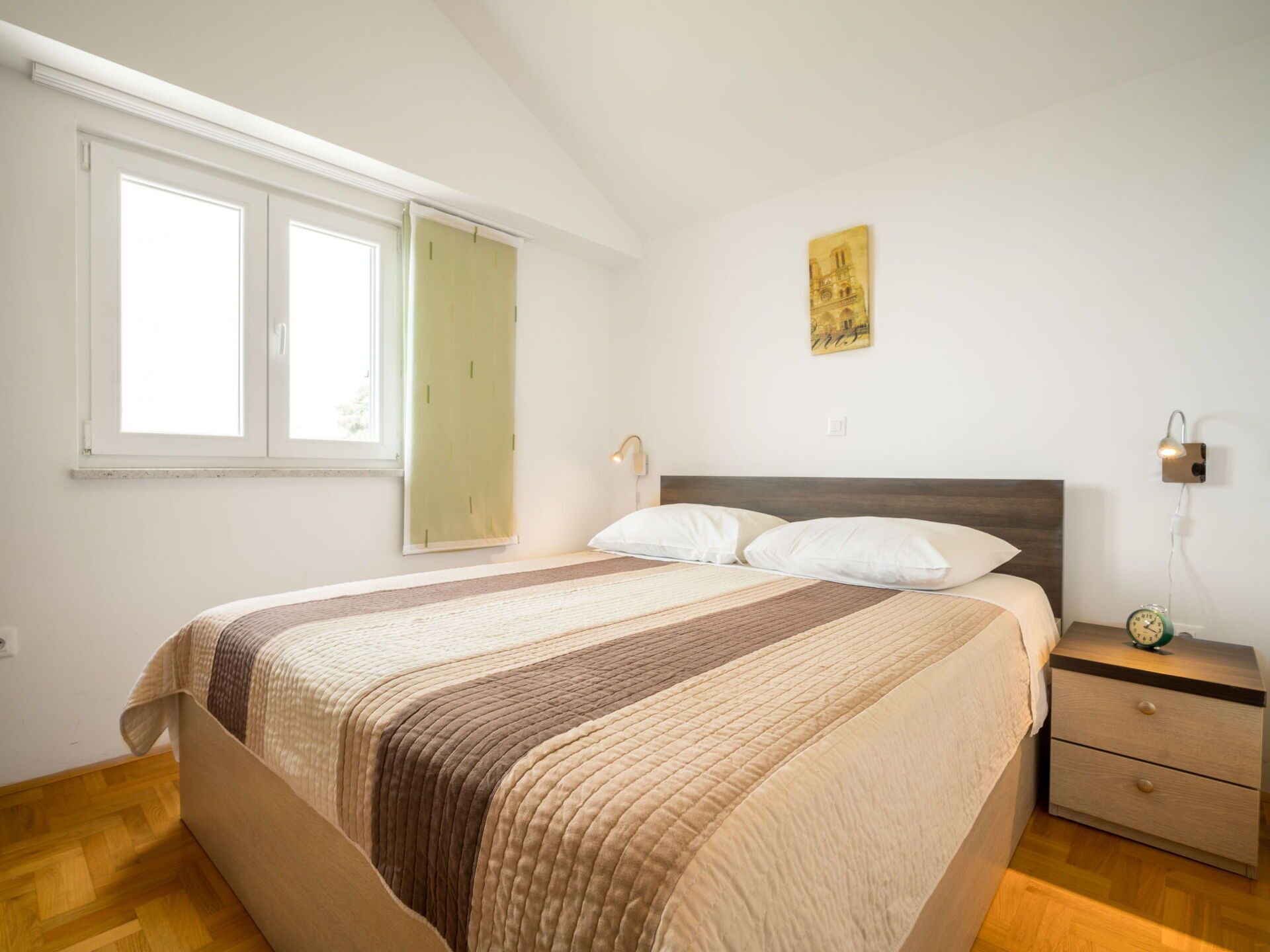 Een slaapkamer met een bed, een nachtkastje, een wandlamp, een schilderij en een raam met gordijn.