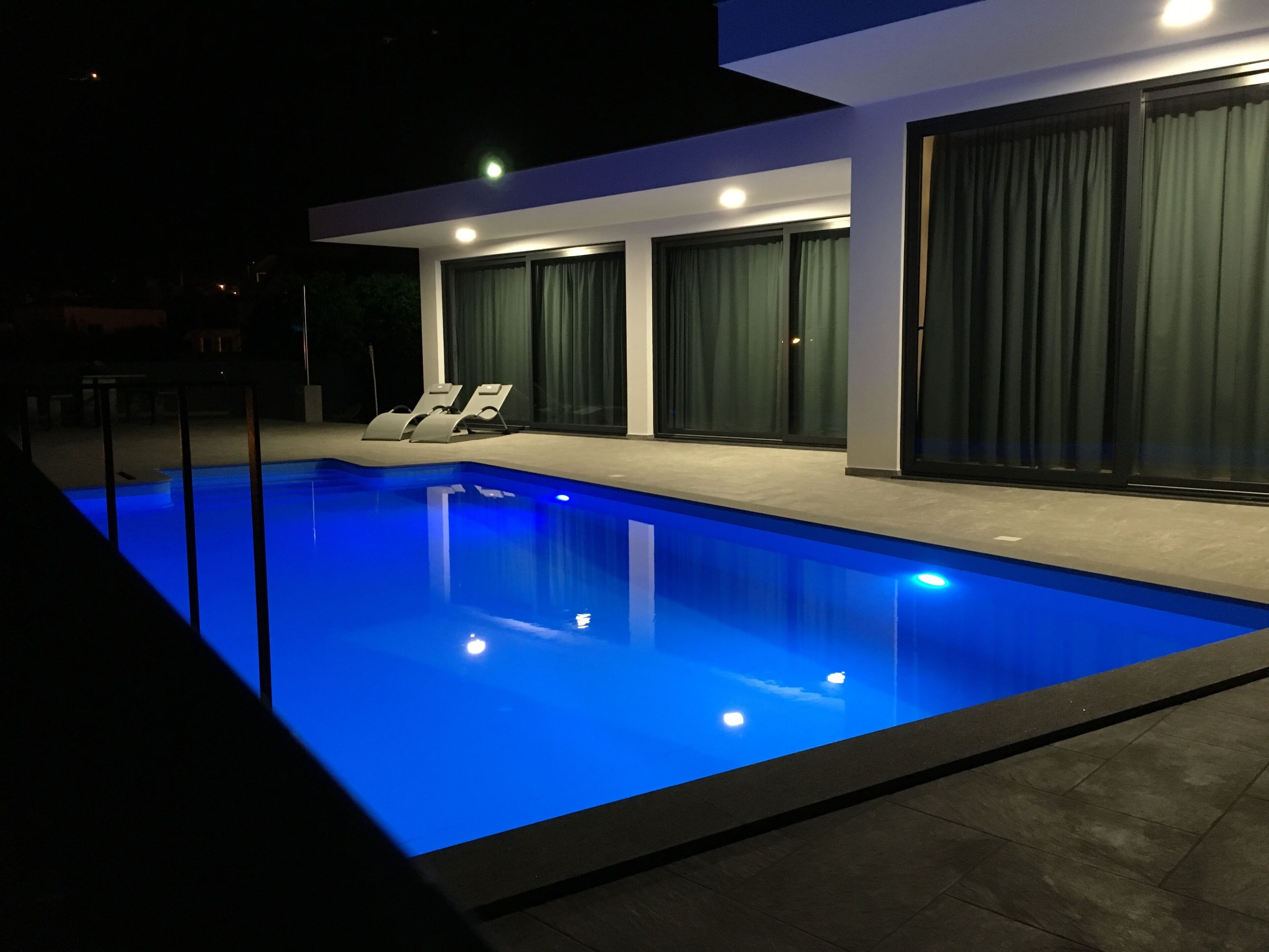 A modern pool area with a clear blue pool, two lounge chairs, and a large sliding glass door.