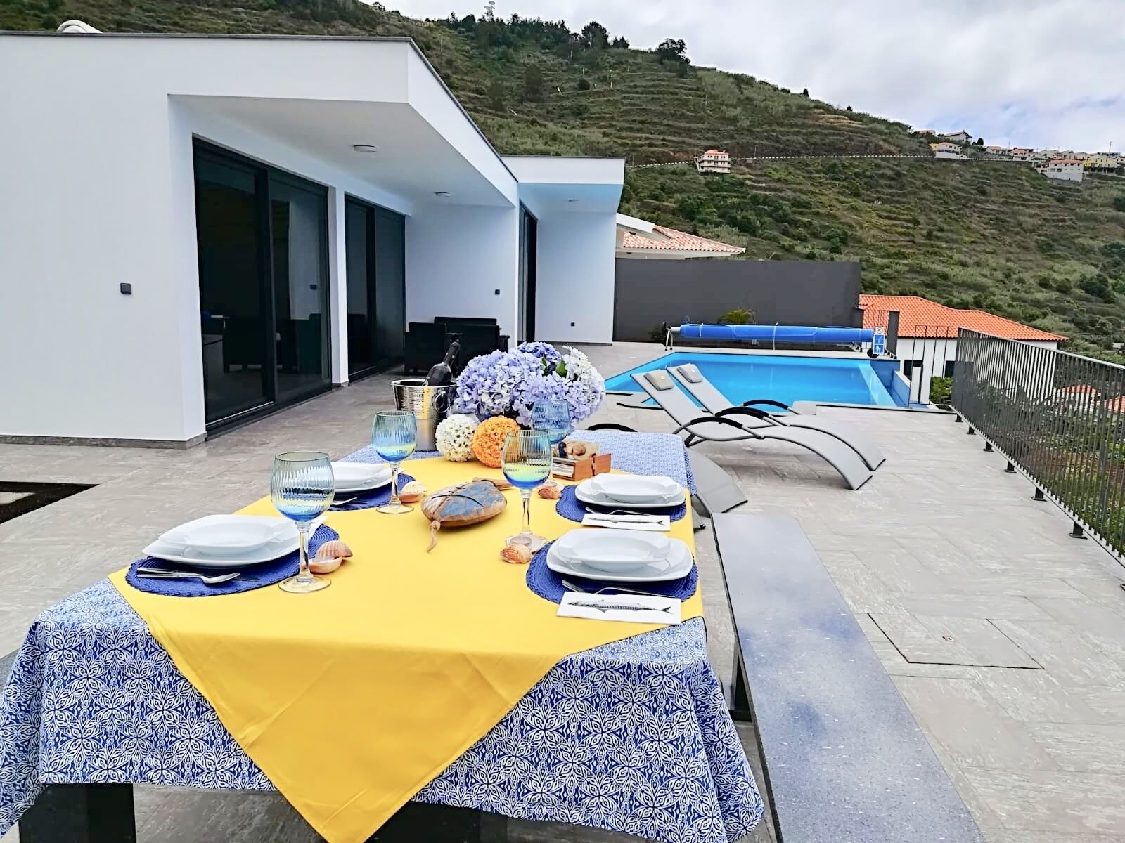 Un tavolo da pranzo apparecchiato con piatti, bicchieri e un centrotavola, con vista su una piscina e una collina.