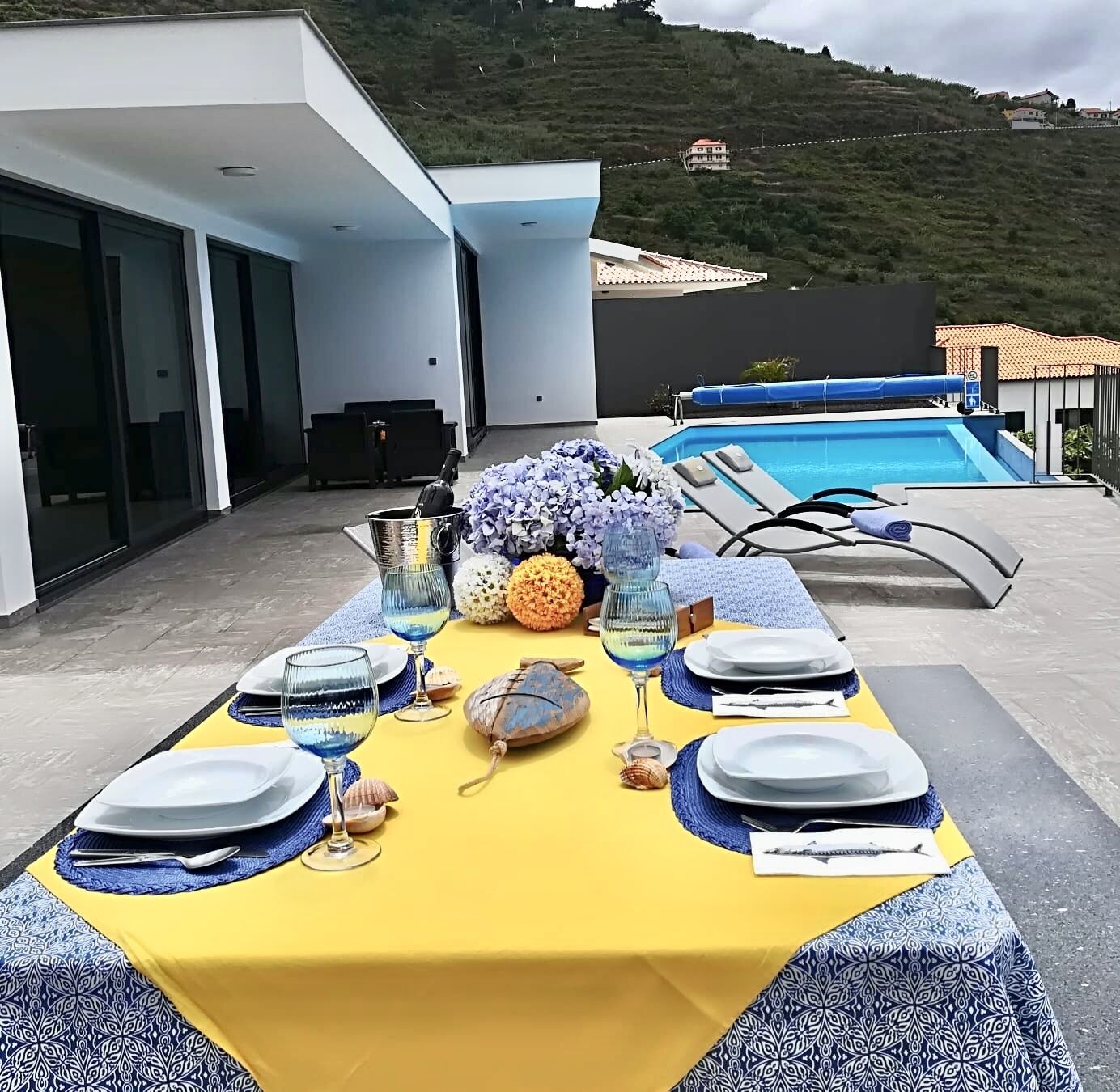 A dining table set with plates, glasses, and decorative items on a yellow tablecloth, overlooking a swimming pool and a hillside.