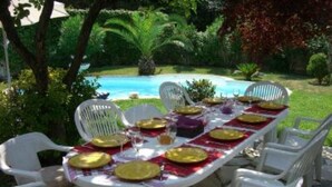 A dining table set outdoors with white chairs, surrounded by lush greenery and a pool. - Single-storey house, rated 4*, air-conditioned, private pool. 10 minutes from the sea. (La Colle-Sur-Loup)