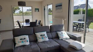 A modern living room with a grey sectional sofa, two pillows with 'HELLO SUMMER' text, a white throw blanket, and a view of an outdoor patio with a table and chairs. - New holiday house 100 meters from the sea (Kerlouan)