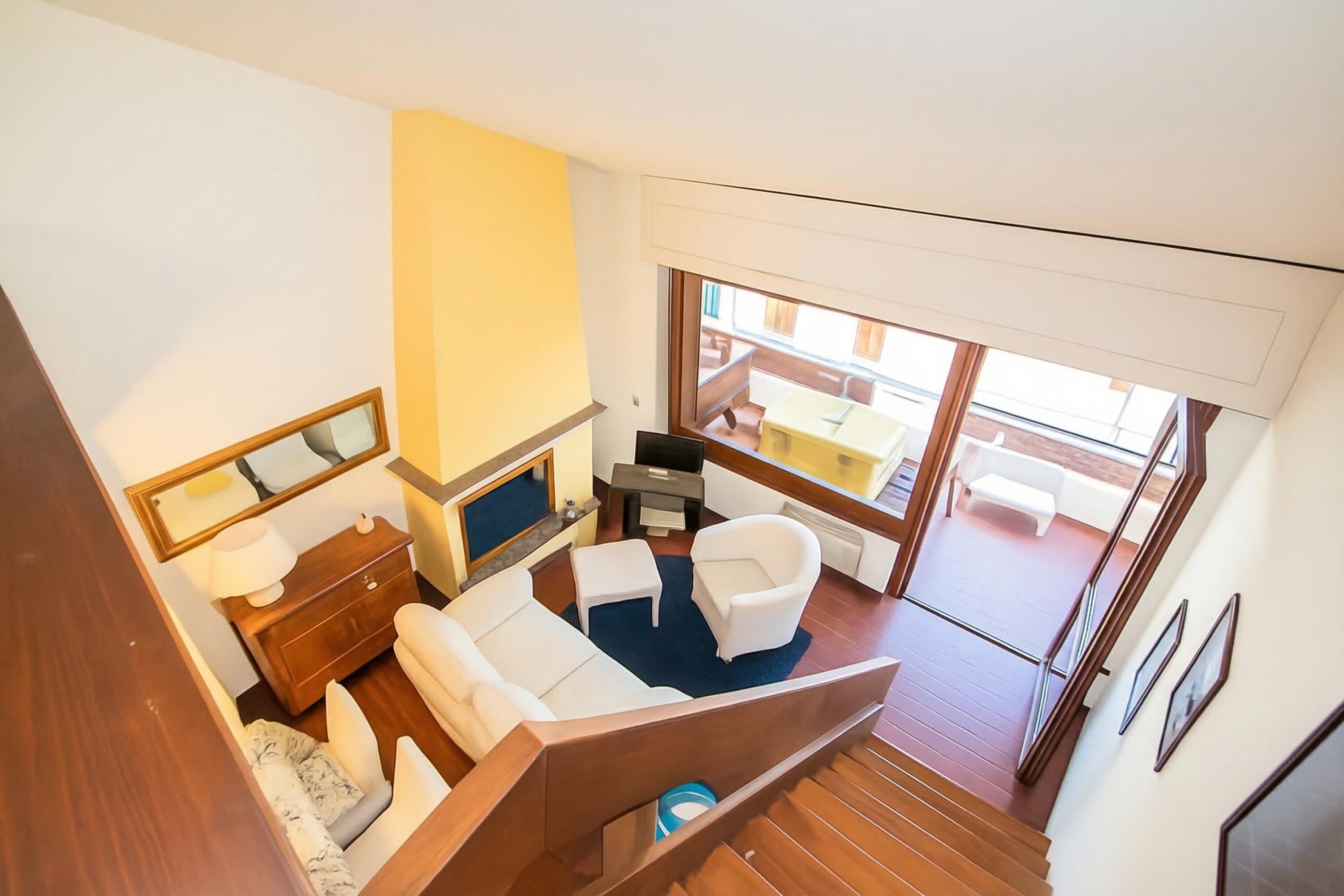 A modern interior with a sofa, armchair, and small table, a wooden staircase, and a view of an outdoor balcony.