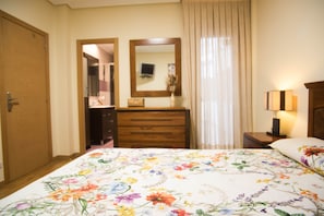 Une chambre à coucher avec une couvre-lit à motifs floraux, une commode en bois, un miroir, une table de chevet avec une lampe et une porte.